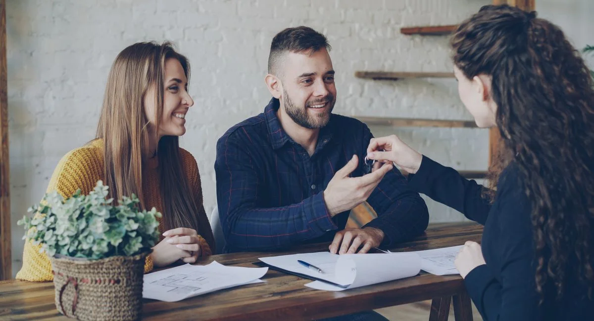 Smiling couple receiving house keys from a female real estate agent at a wooden desk.
