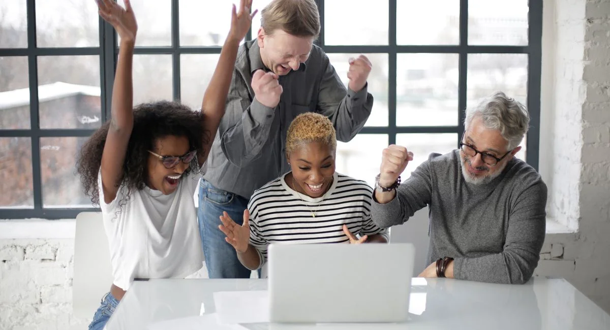 Cheerful multiethnic coworkers of different ages in casual clothing clenching fists and raising hands up in joy during succes
