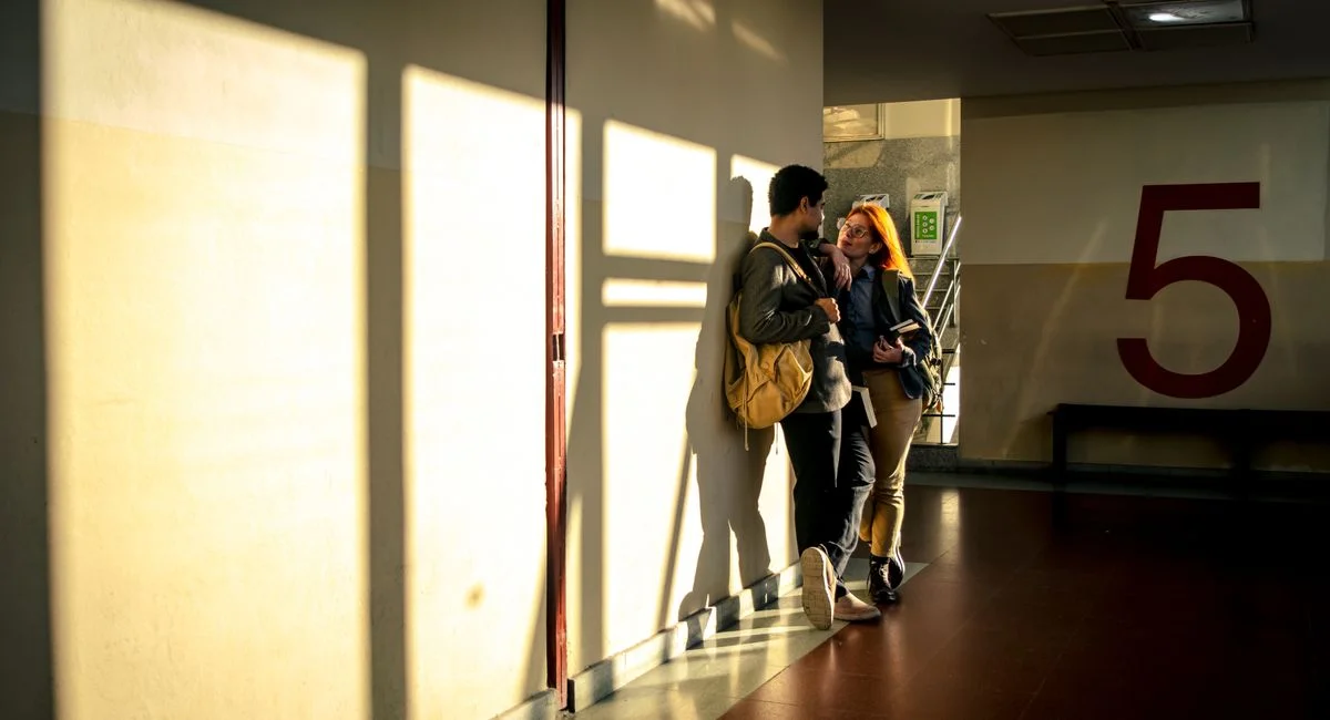 Two students conversing in a sunlit hallway, casting long shadows on a wall with a large number 5.