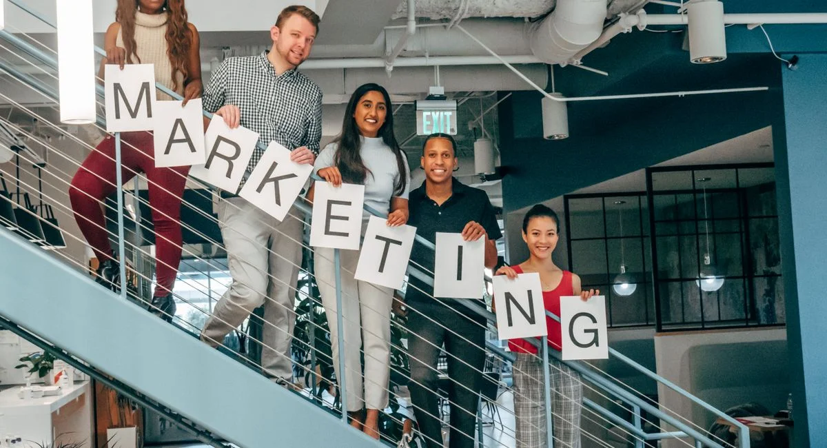 Five colleagues smiling and holding a marketing sign on an office staircase, showcasing teamwork.