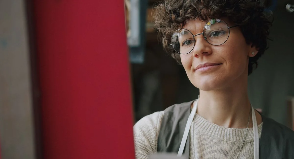 A young woman artist wearing glasses paints attentively on a canvas indoors.